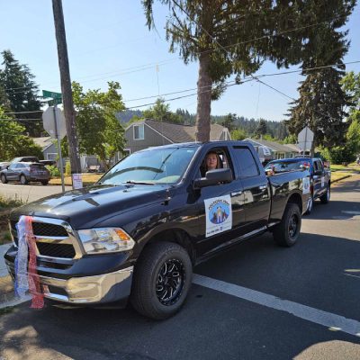 A truck adorned with a red ribbon drives along a city street, showcasing a festive and cheerful atmosphere A blue truck parked with a man seated in the driver's seat, ready for the journey ahead